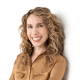 A person with long curly hair and a warm smile, wearing a brown blouse, stands against a white background.